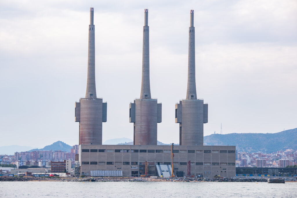 A large waterfront power plant with three tall chimneys and a rectangular base stands against the cityscape and hills under a cloudy sky, setting an industrial scene reminiscent of installations showcased at 3daysofdesign.