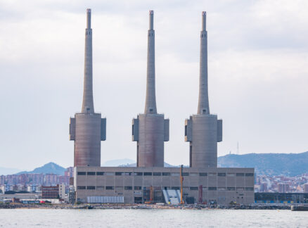 A large waterfront power plant with three tall chimneys and a rectangular base stands against the cityscape and hills under a cloudy sky, setting an industrial scene reminiscent of installations showcased at 3daysofdesign.