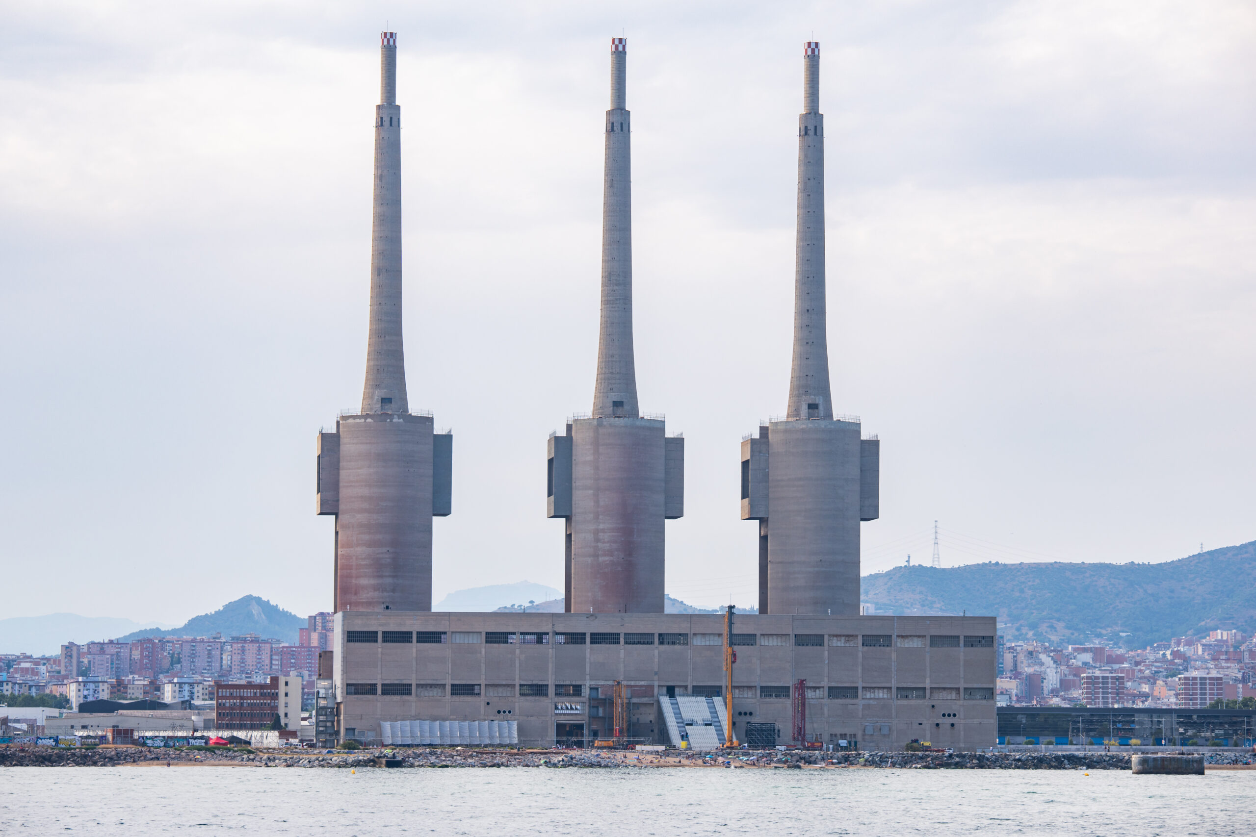 A large waterfront power plant with three tall chimneys and a rectangular base stands against the cityscape and hills under a cloudy sky, setting an industrial scene reminiscent of installations showcased at 3daysofdesign.