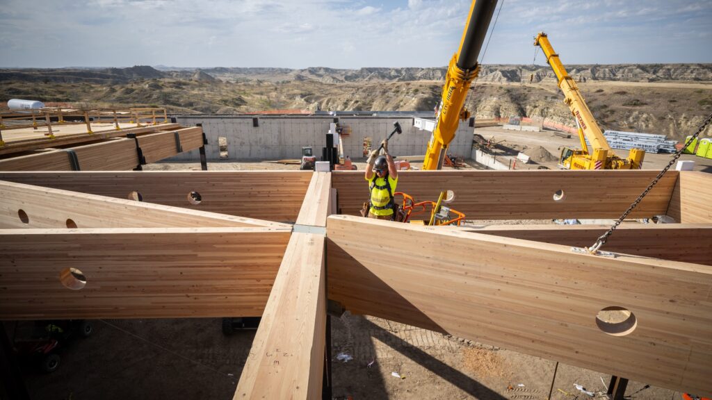 Construction worker in a safety vest operates a crane to position large wooden beams and other materials at a building site in a desert landscape under a partly cloudy sky.