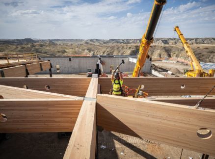 Construction workers assemble large wooden beams with cranes at a building site in a dry, hilly landscape under a partly cloudy sky, where the design subtly incorporates biophilia to harmonize the structure with its natural surroundings.