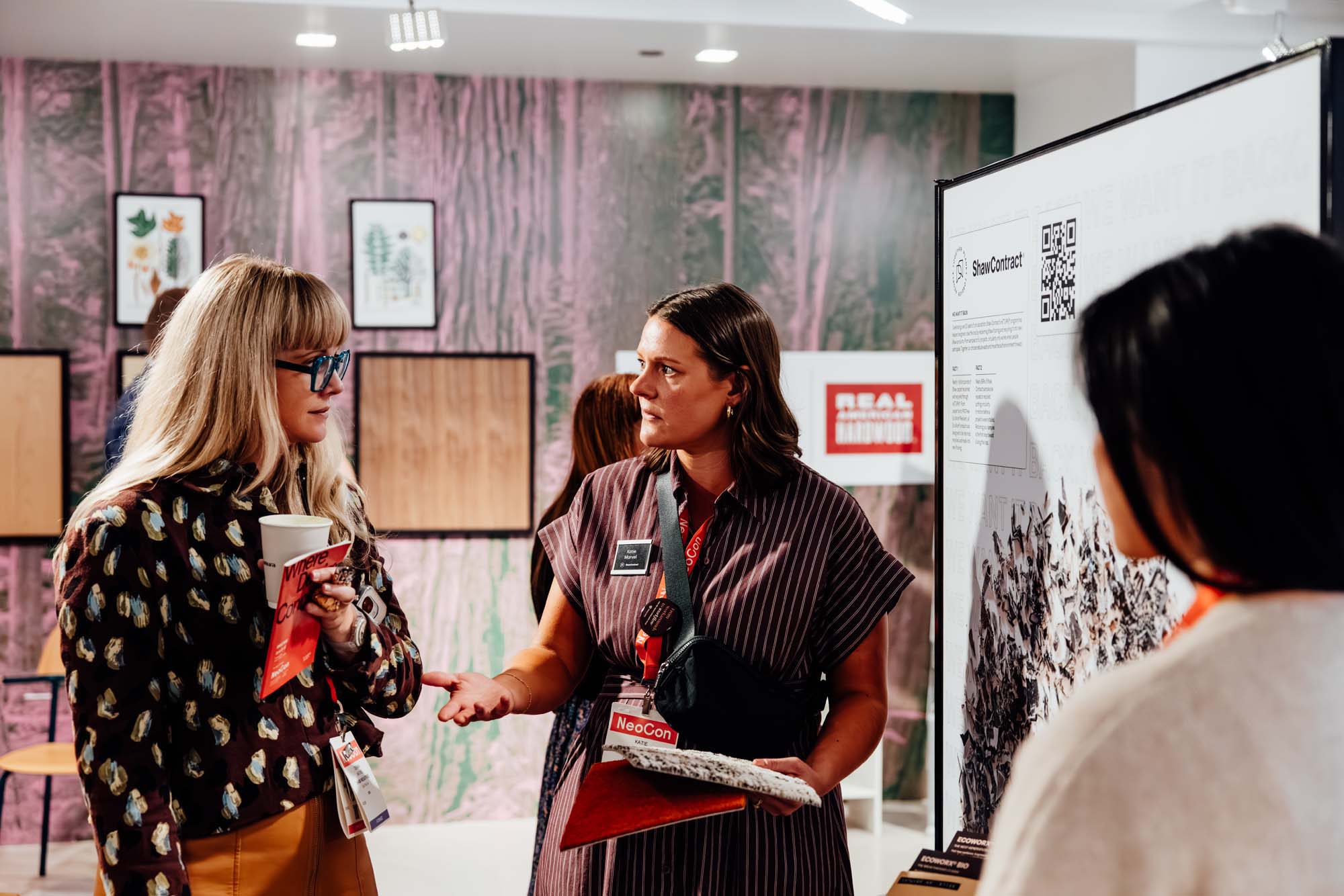 Three women stand and converse near a METROPOLIS Sustainability Lab display board filled with charts and QR codes in a modern room with patterned walls and framed artwork.