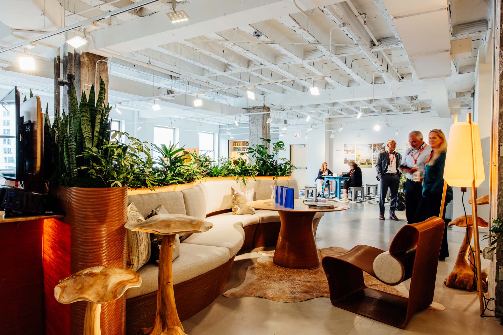 Modern office lounge with wooden furniture, potted plants, and natural light. Three people stand talking in the background near desks and windows of the METROPOLIS Sustainability Lab.