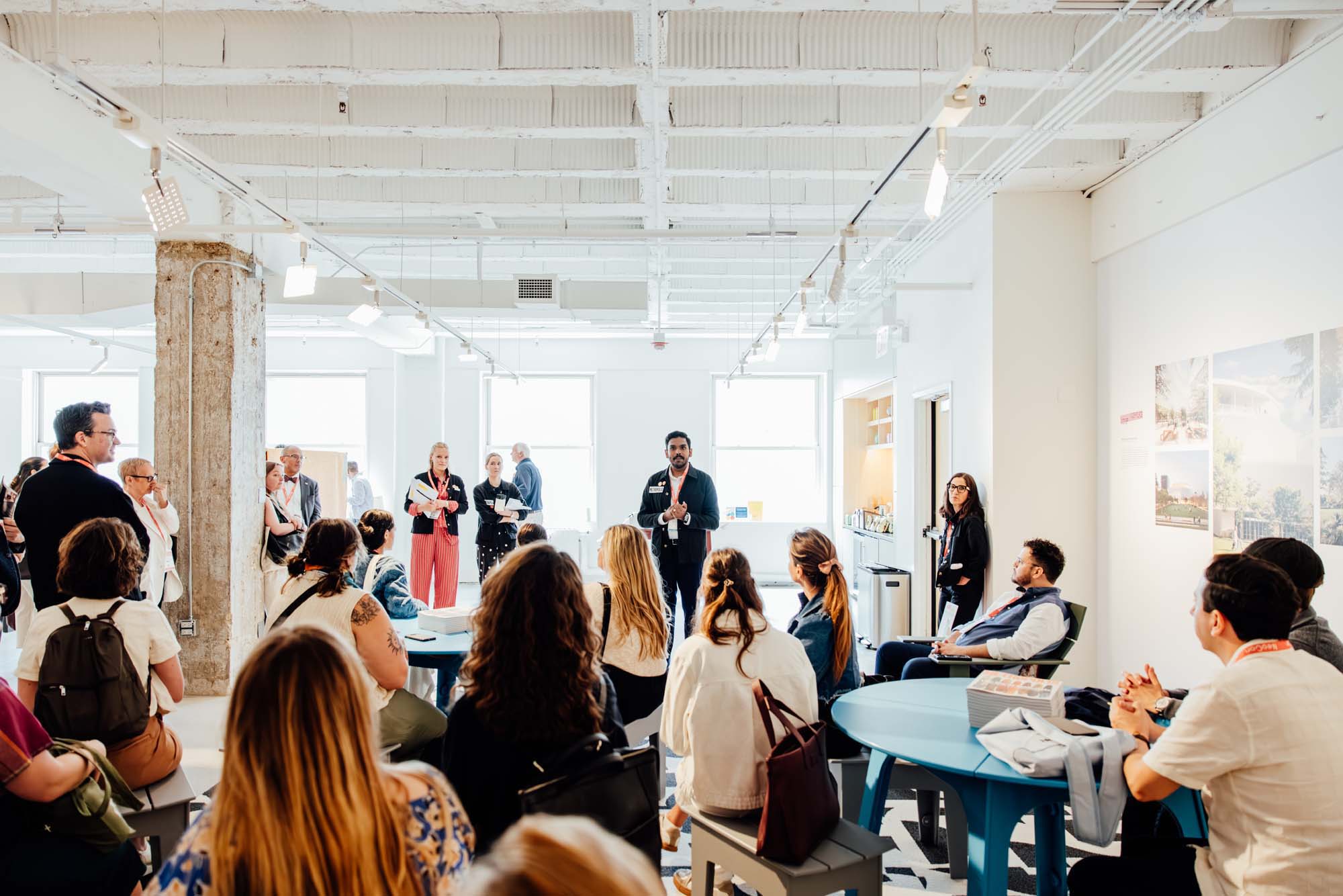 A group of people sits and stands in a modern, well-lit room at the METROPOLIS Sustainability Lab, listening to a person speaking at the front near a column.