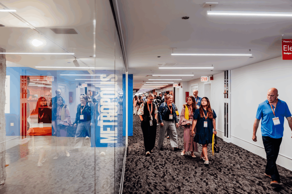 A group of people with name badges walk down a modern hallway with glass walls and visible "METROPOLIS" signage, likely attending the METROPOLIS Leadership Summit, while others are reflected in the glass.
