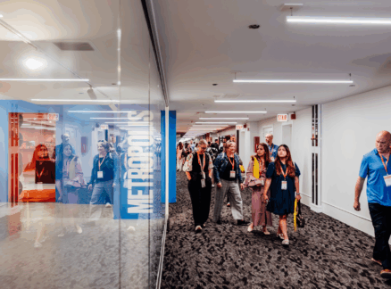 A group of people with name badges walk down a modern hallway with glass walls and visible "METROPOLIS" signage, likely attending the METROPOLIS Leadership Summit, while others are reflected in the glass.