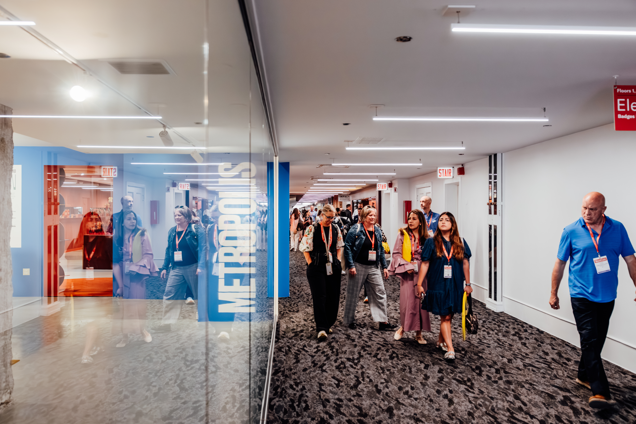 A group of people with name badges walk down a modern hallway with glass walls and visible "METROPOLIS" signage, likely attending the METROPOLIS Leadership Summit, while others are reflected in the glass.