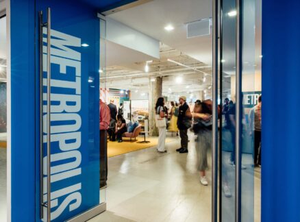 People gather and converse inside a modern, brightly lit space, viewed through open glass doors adorned with "METROPOLIS Sustainability Lab" signage.