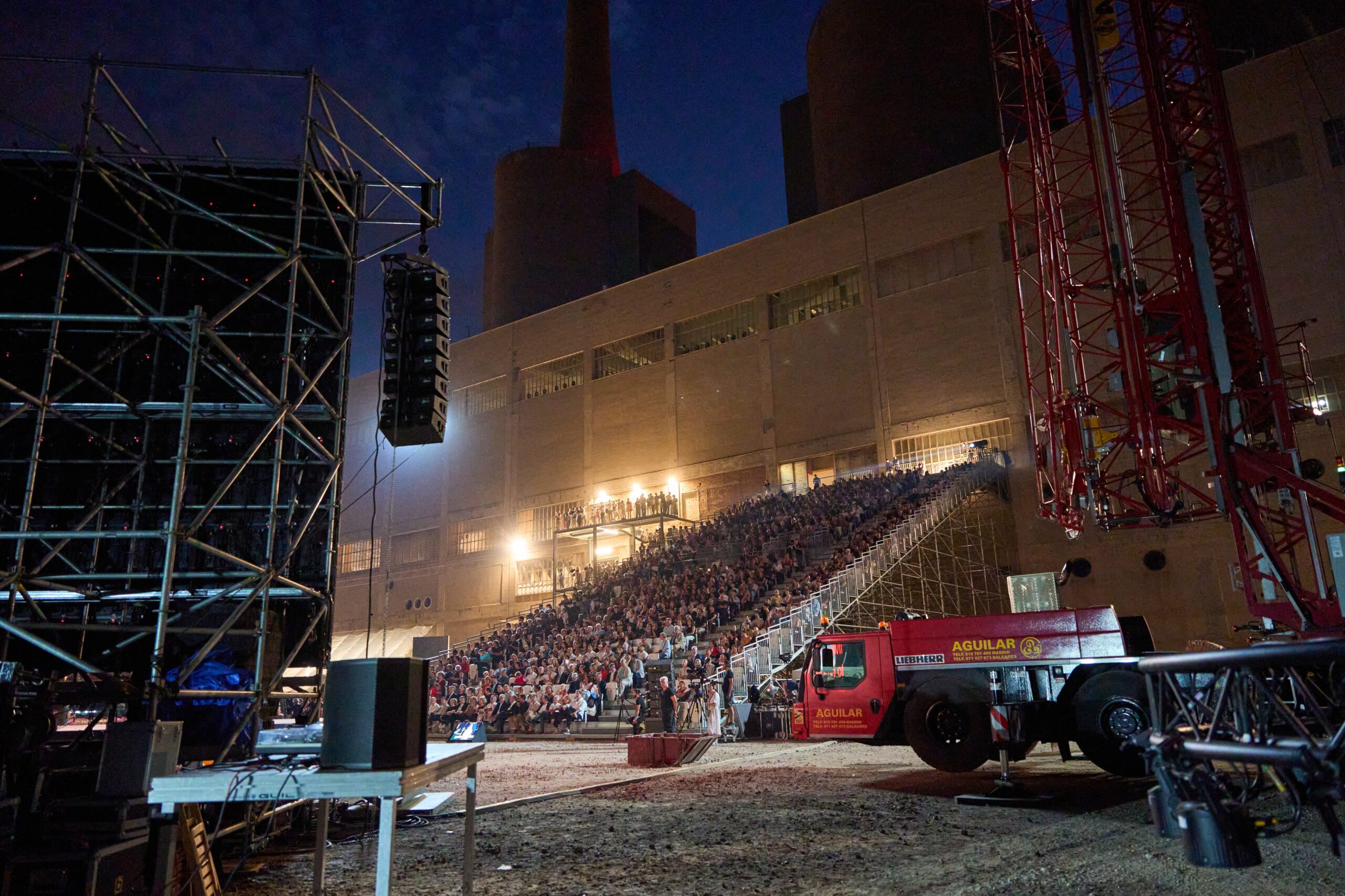 An audience sits in bleachers outside a large industrial building at night during 3daysofdesign, with stage equipment, scaffolding, and a red truck visible in the foreground.