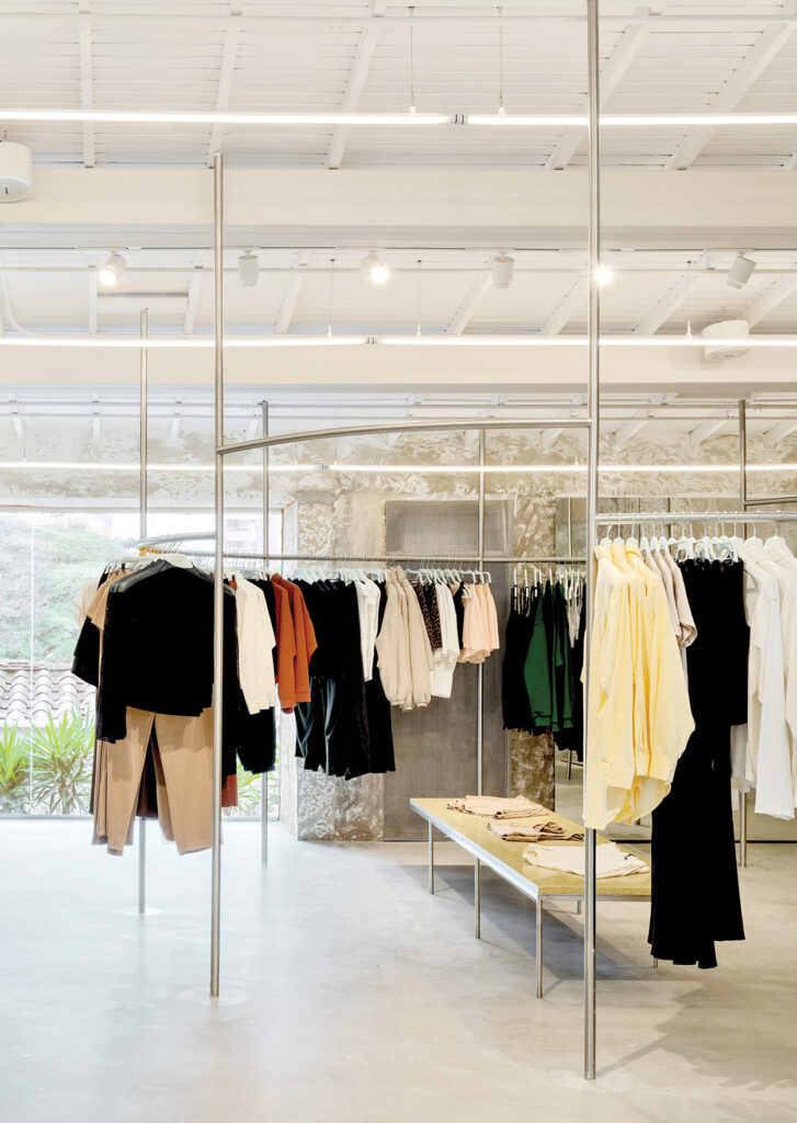 Minimalist clothing store interior by Latin Matter with metal racks showcasing assorted shirts, pants, and jackets. A central table displays folded items. Light-colored floor and ceiling with ample natural light create an inviting atmosphere.
