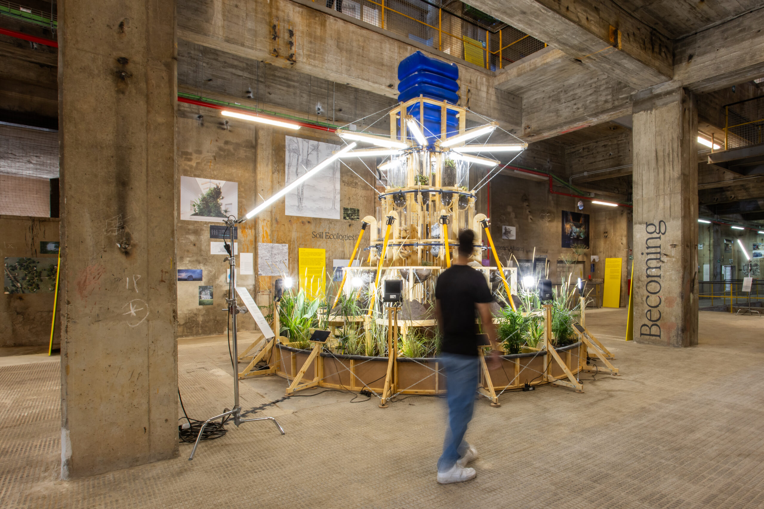 A person walks past a large, tiered wooden structure with plants and fluorescent lights at 3daysofdesign, set in an industrial indoor space with concrete pillars and striking art installations.