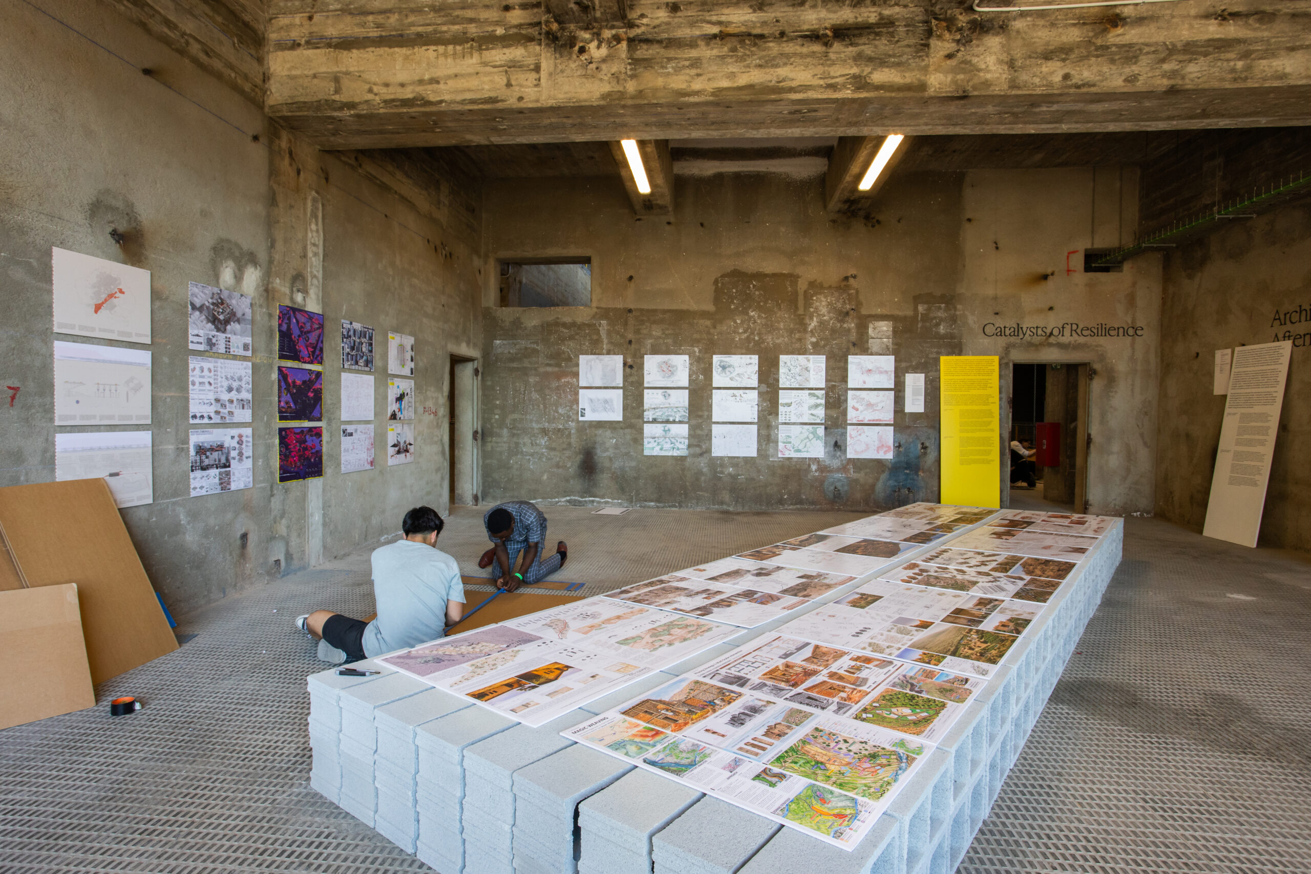 Two people work on the floor of an industrial-style room during 3daysofdesign, displaying architectural plans and drawings on cinder blocks and walls under fluorescent lights.
