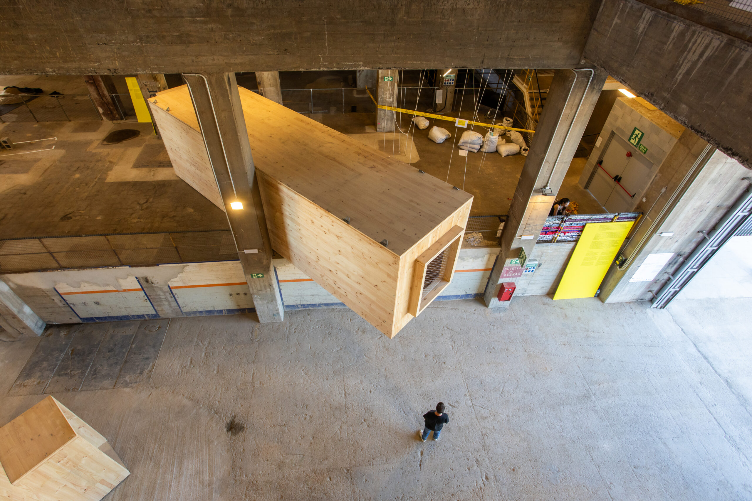 A person stands below a large wooden structure suspended in an industrial building with concrete walls and yellow doors, capturing the creative spirit of 3daysofdesign.