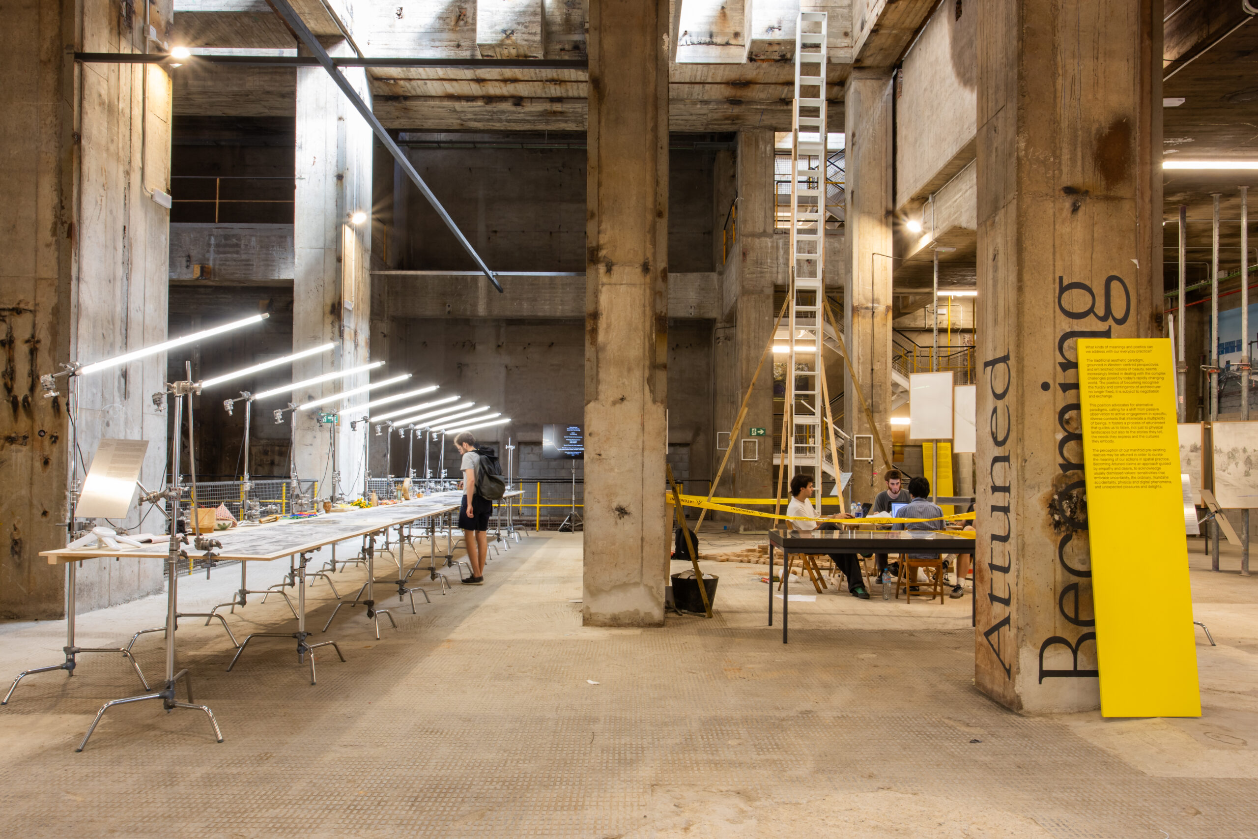 An industrial space with concrete pillars features long tables with lights, people working at a desk, and a large yellow sign reading "Attuned Becoming"—a creative hub showcased during 3daysofdesign.