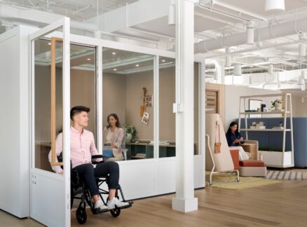 A man in a wheelchair leaves a glass office, while two women work at separate desks in a modern, neuroinclusive open-plan workspace.