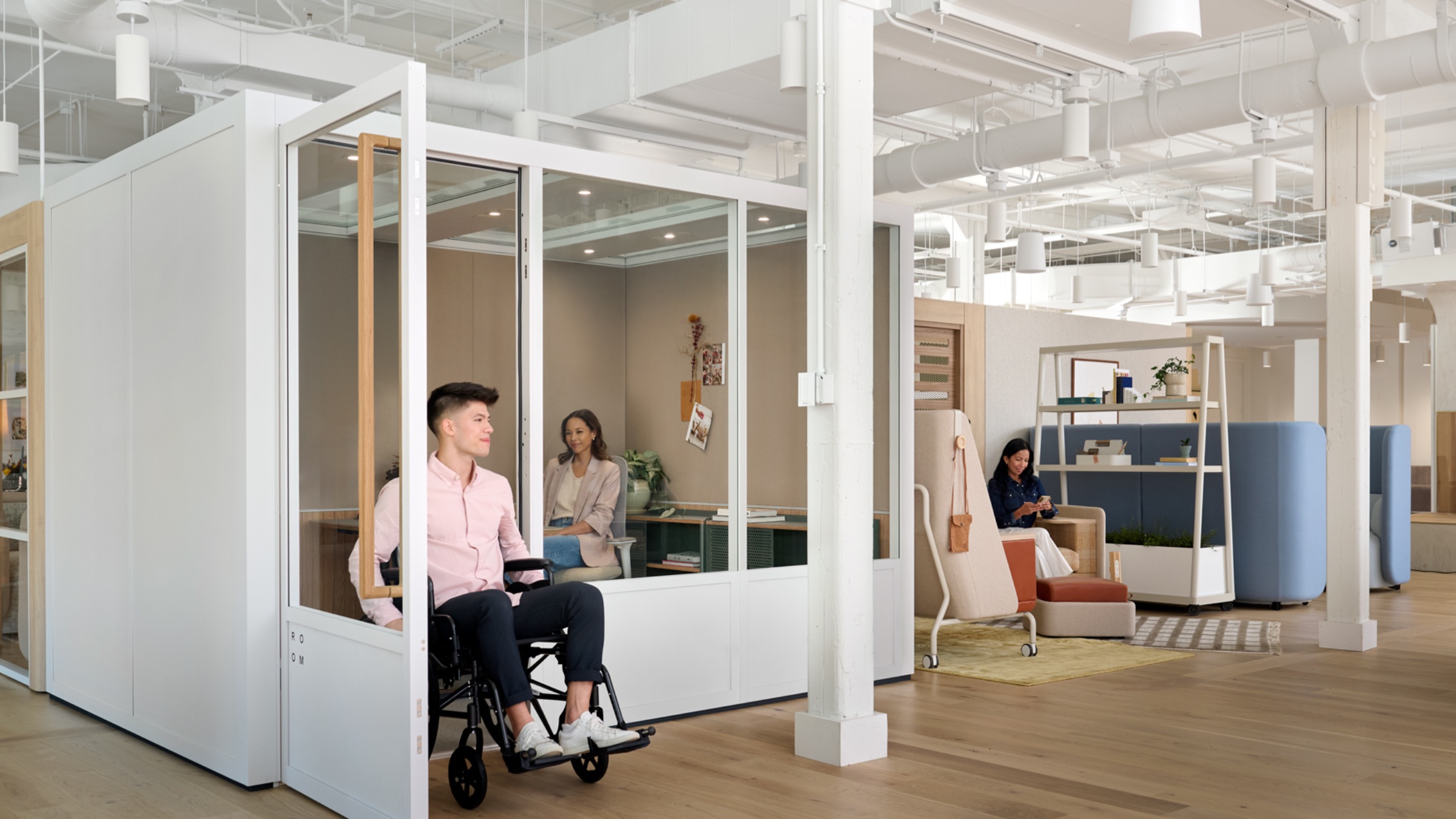 A man in a wheelchair leaves a glass office, while two women work at separate desks in a modern, neuroinclusive open-plan workspace.