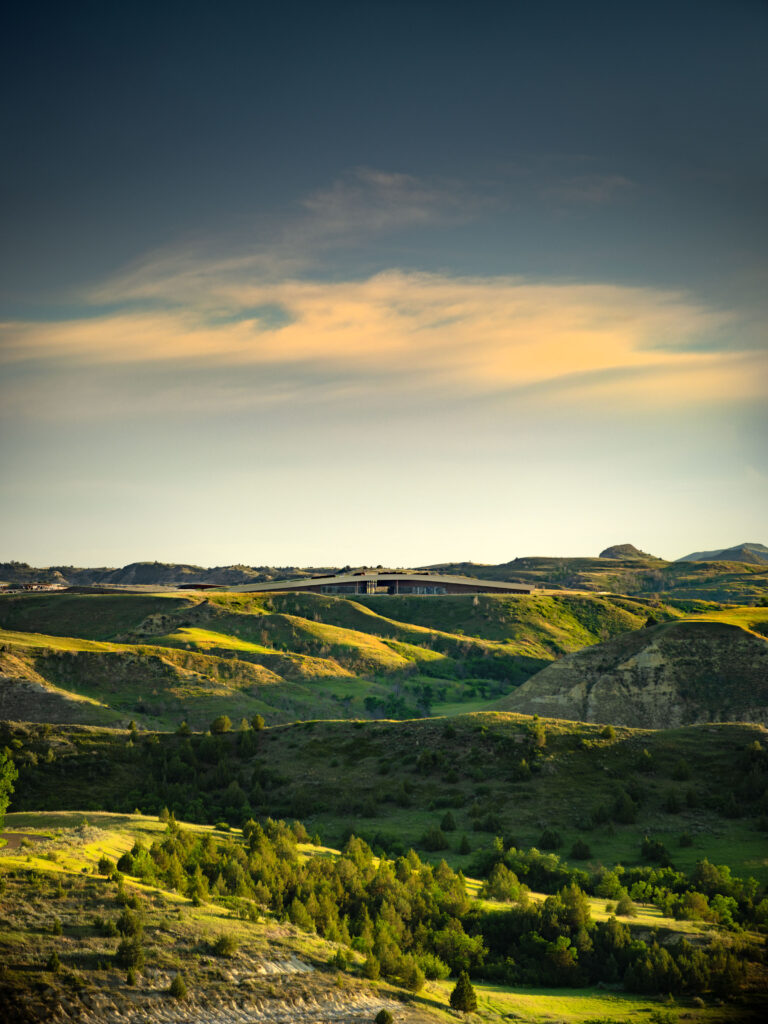 Rolling hills with scattered green trees under a blue sky with yellow clouds during daylight, near the future site of the Theodore Roosevelt Presidential Library.