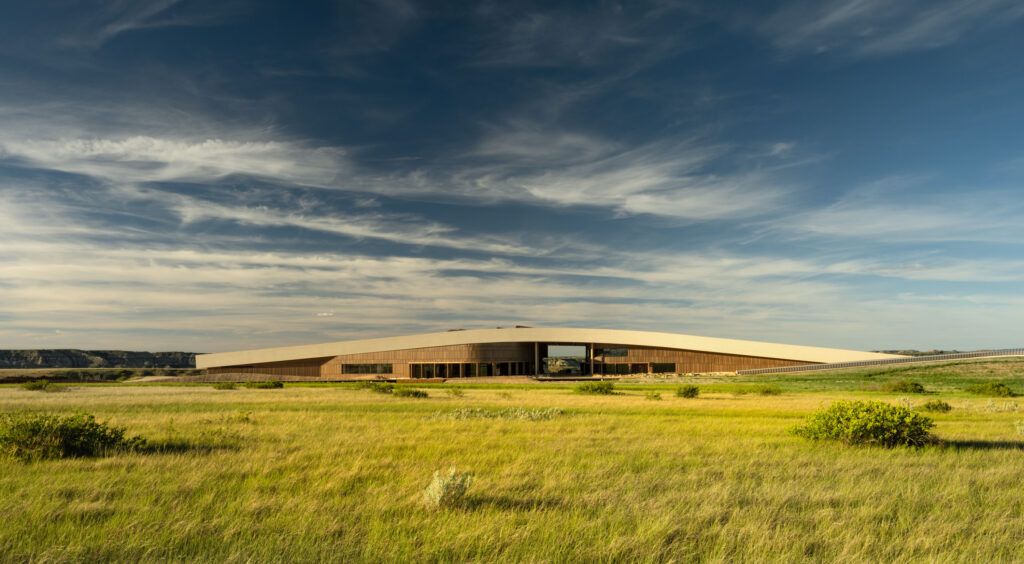 A modern, low-profile building with a wooden exterior, designed for the Theodore Roosevelt Presidential Library, sits on a grassy plain under a blue sky with wispy clouds.