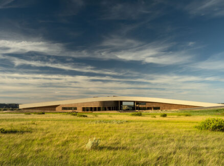A modern, low-profile building with a wooden exterior, designed for the Theodore Roosevelt Presidential Library, sits on a grassy plain under a blue sky with wispy clouds.