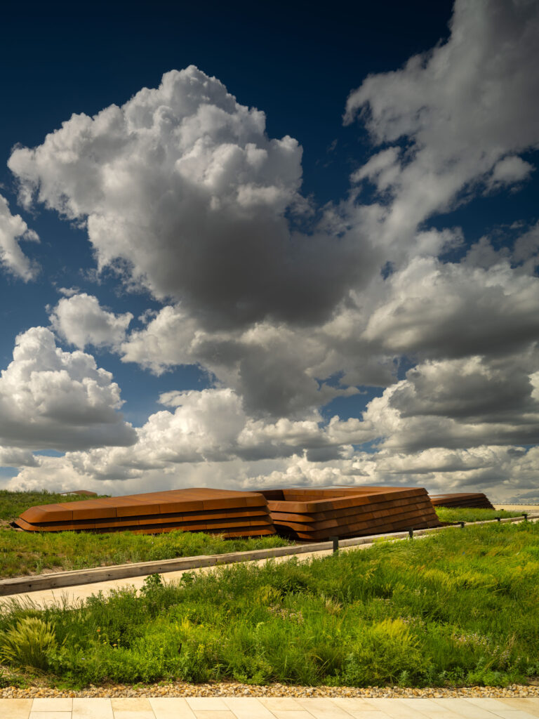 Modern low-profile building with a rust-colored exterior, home to the Theodore Roosevelt Presidential Library, set amid green vegetation under a dramatic sky with large, fluffy clouds.