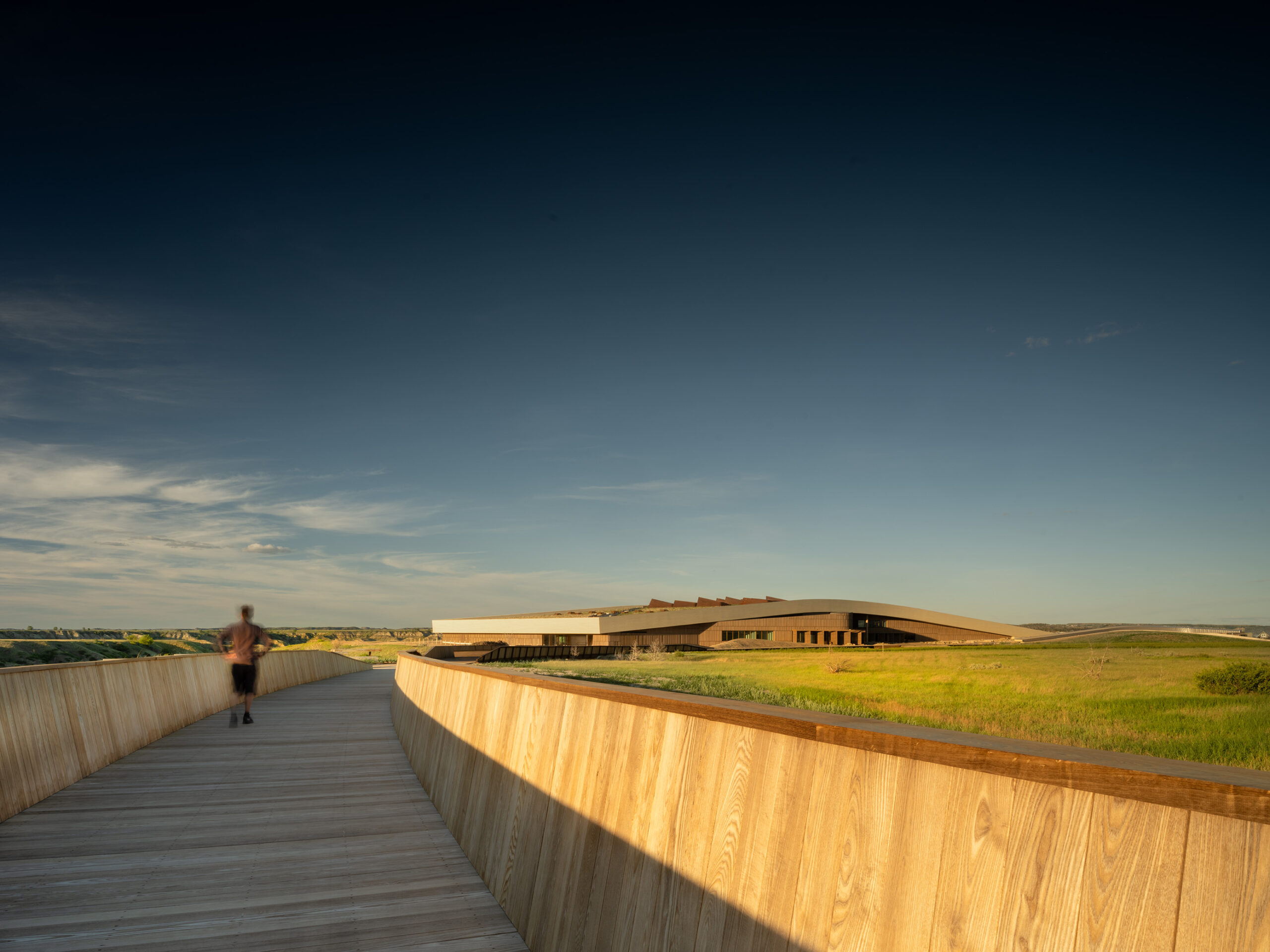 A person walks on a wide wooden pathway toward the Theodore Roosevelt Presidential Library, set in an open grassy landscape under a clear sky.