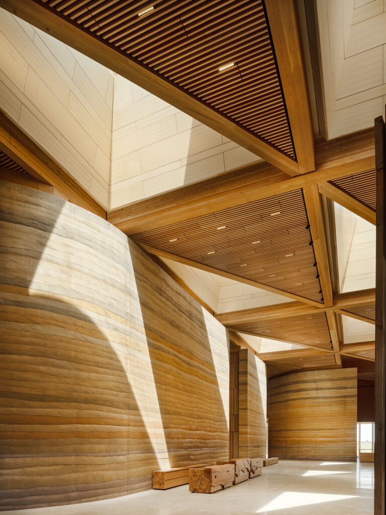 Modern interior of the Theodore Roosevelt Presidential Library with large wooden beams, ribbed wood ceiling, curved rammed-earth walls, and natural light streaming through skylights; wooden benches line the space.