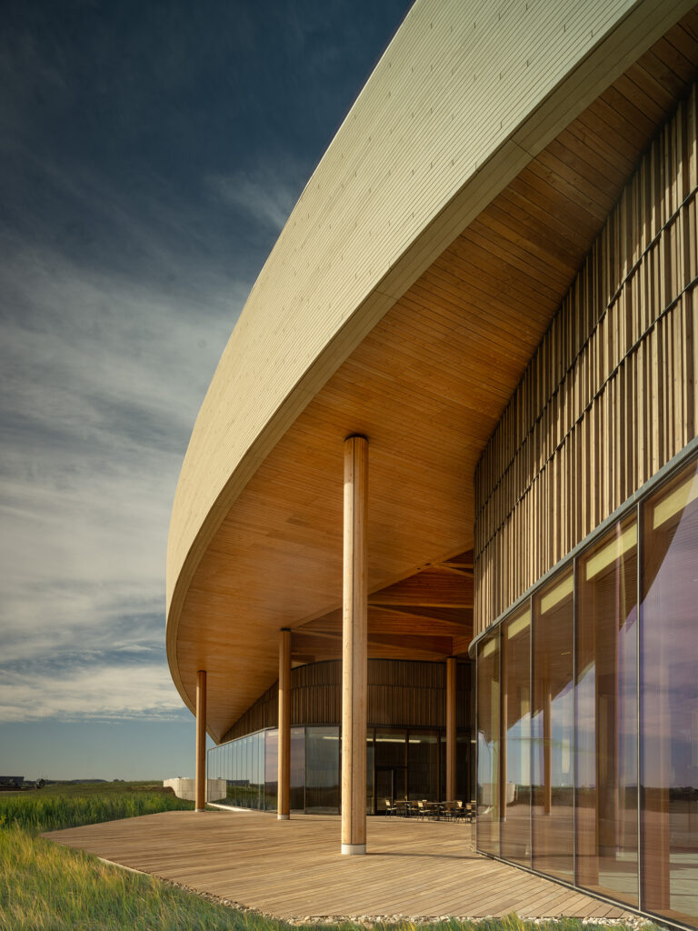 Modern building with a curved wooden roof, glass walls, and wooden pillars, set on a wooden deck under a partly cloudy sky—an architectural gem reminiscent of the Theodore Roosevelt Presidential Library.