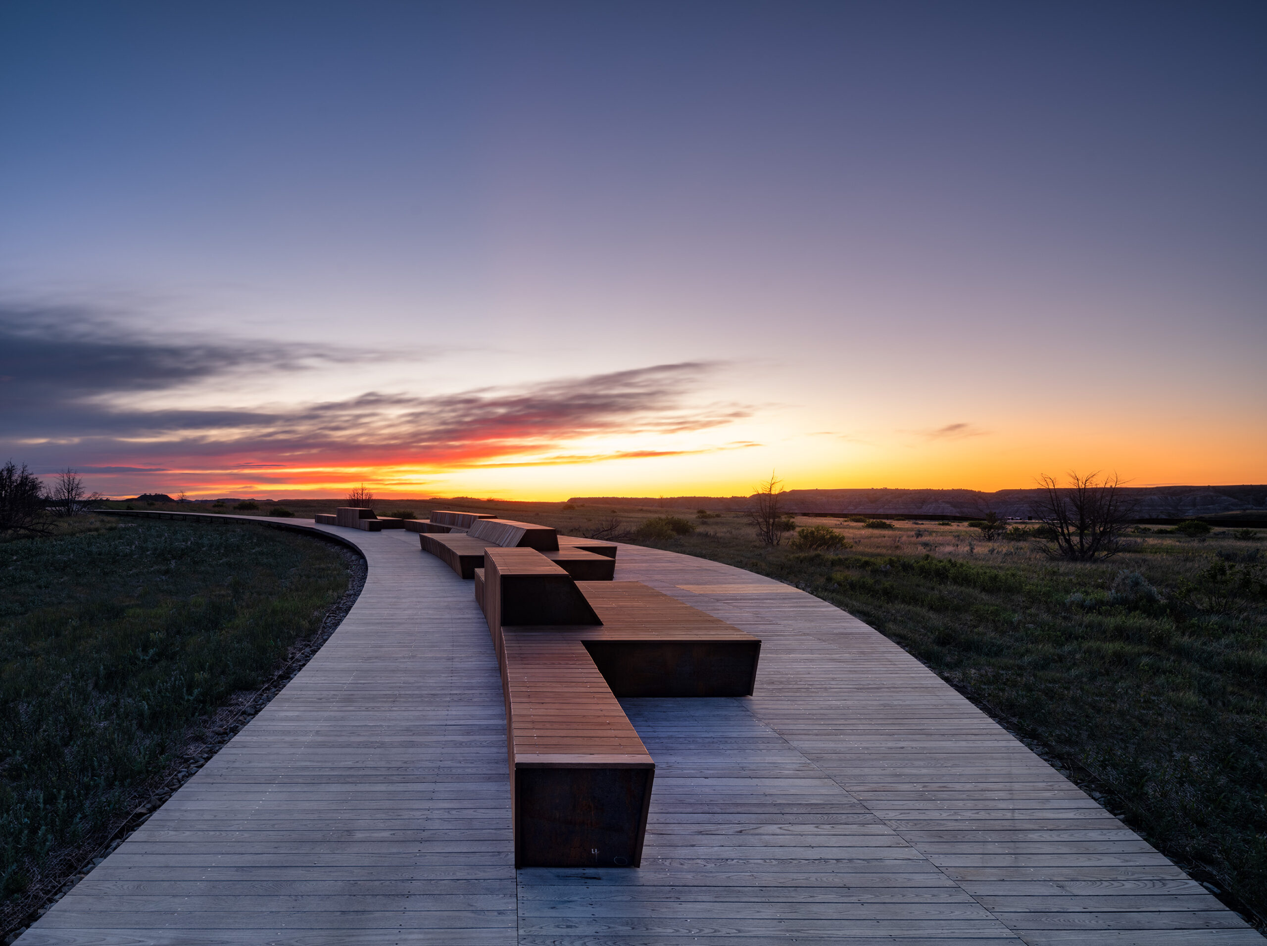 A curved boardwalk with geometric benches runs through grassy fields at sunset near the Theodore Roosevelt Presidential Library, beneath a colorful sky with scattered clouds.