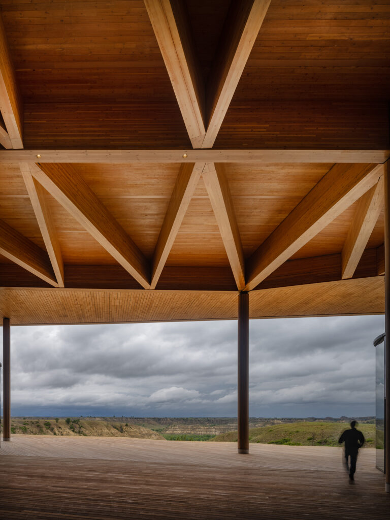 A person walks under a large wooden canopy with exposed beams at the Theodore Roosevelt Presidential Library, overlooking a landscape of hills beneath a cloudy sky.