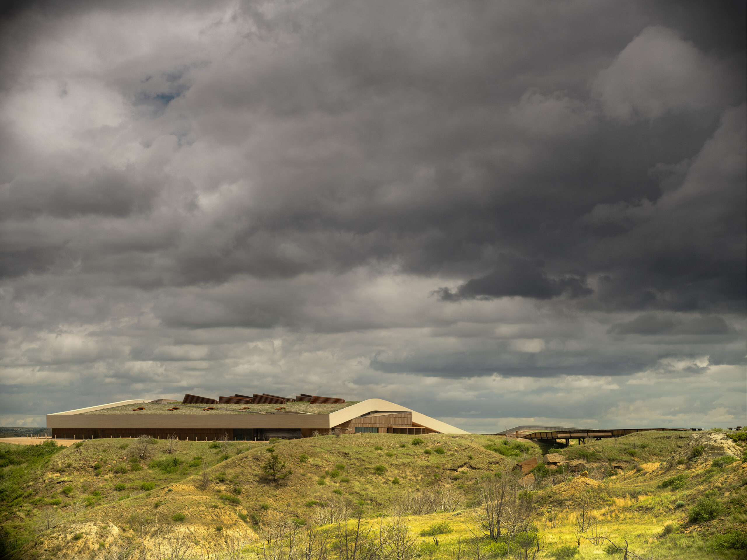 A modern, low-profile building with angular roofs sits on a grassy hill under a dramatic, cloudy sky—the future home of the Theodore Roosevelt Presidential Library.