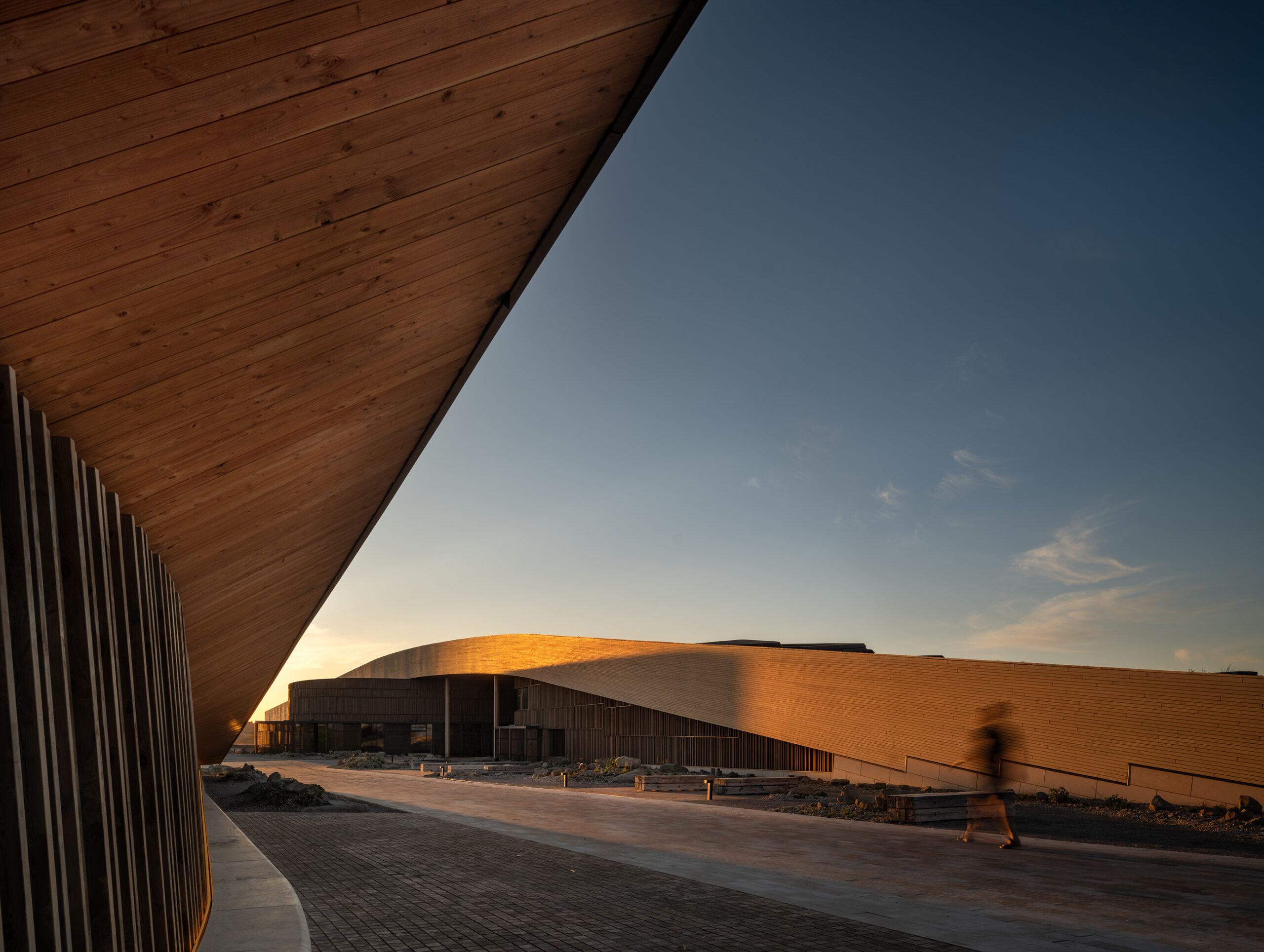 Contemporary building with a curved wooden facade at sunset, casting long shadows at the Theodore Roosevelt Presidential Library, with a blurred figure walking on the paved path in the foreground.