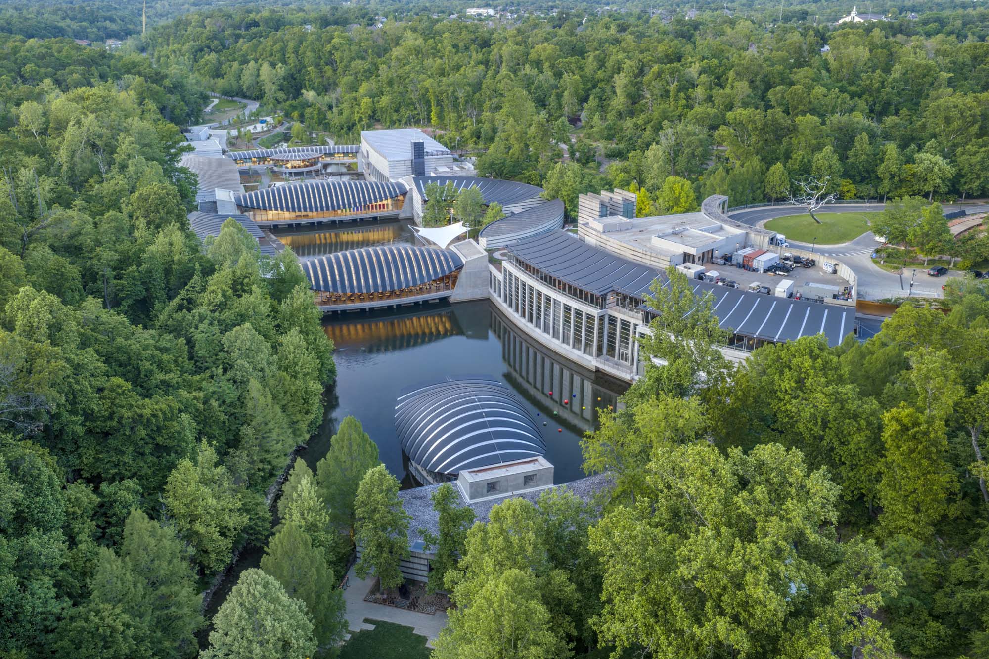 A modern art museum complex with curved glass and metal roofs sits among dense green trees, surrounded by water features and walking paths.