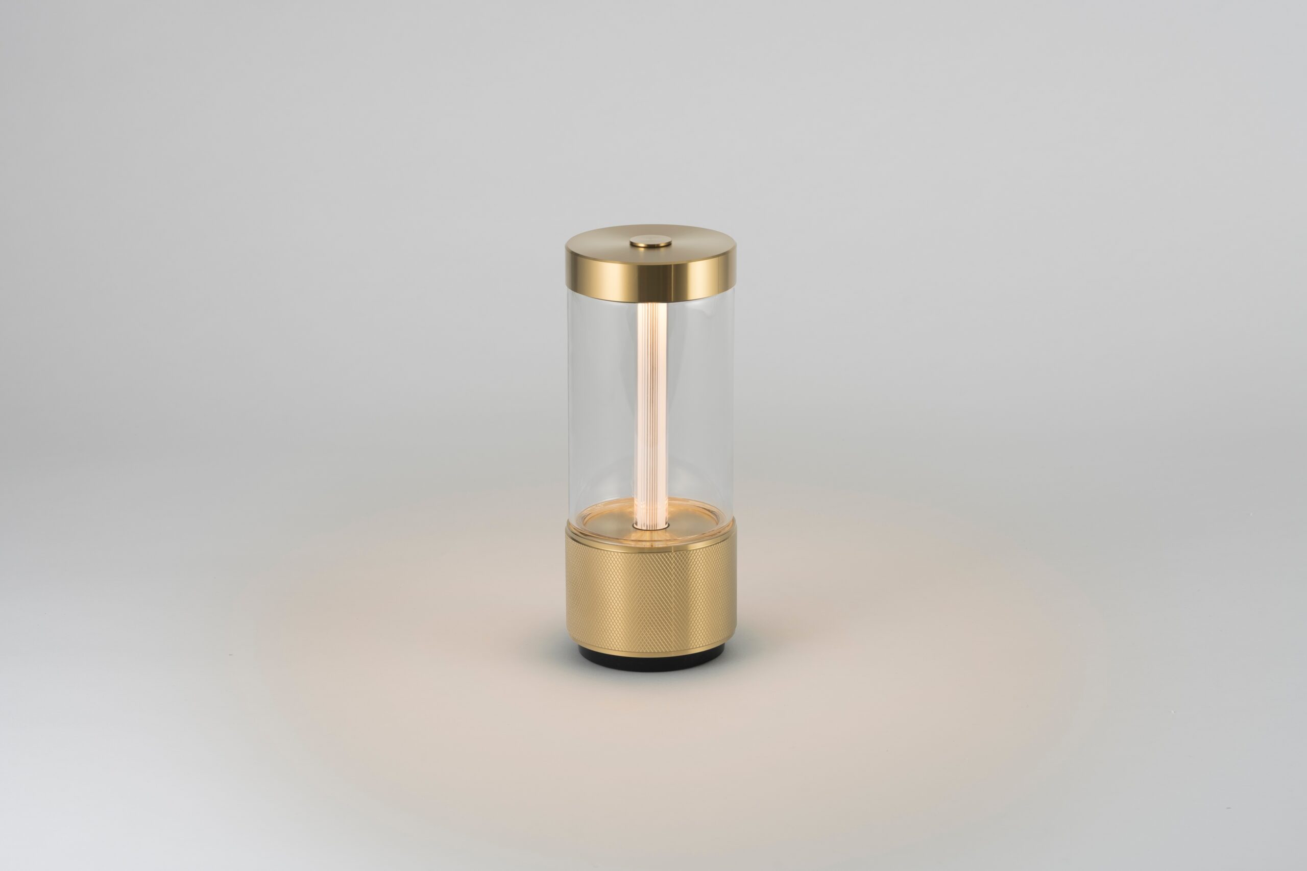 A cylindrical lamp with a gold base and top, featuring a clear glass body and a vertical light tube in the center, placed on a plain light gray background.