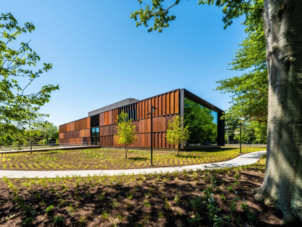 Modern building with rust-colored panels and glass walls, surrounded by young trees, landscaped grounds, and a clear blue sky.