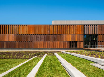 Modern building with rust-colored metal panels and horizontal windows, surrounded by green lawn, concrete steps, and clear blue sky—a scene that echoes the timeless spirit of Bruce Springsteen’s Americana.
