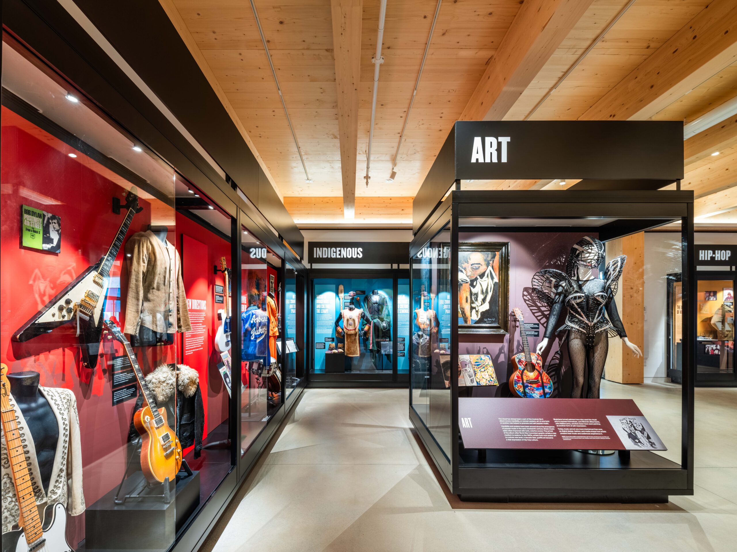 A modern museum exhibit displays guitars, costumes, and artifacts in glass cases labeled with genres like Art, Indigenous, and Hip-Hop under wooden ceilings.