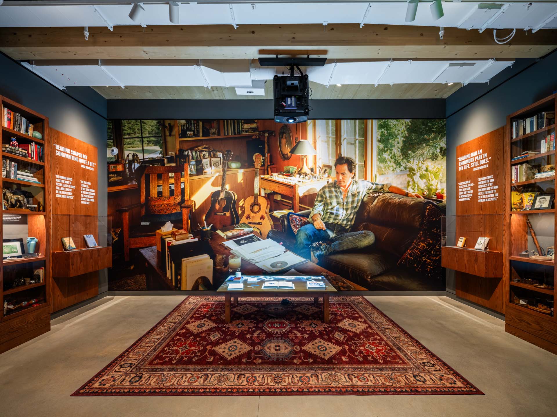 A museum exhibit features a large wall photo of a man in a cozy room with guitars, books, and papers, surrounded by display cases of memorabilia and a patterned rug on the floor.