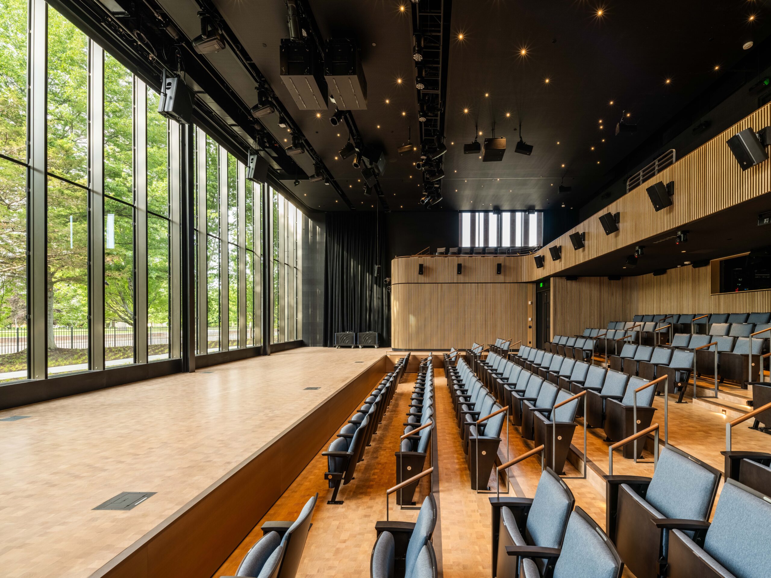 Modern auditorium with tiered seating, large windows on one side, a wooden stage, and black ceiling with lighting fixtures. Natural light enters from the left side through tall glass panels.