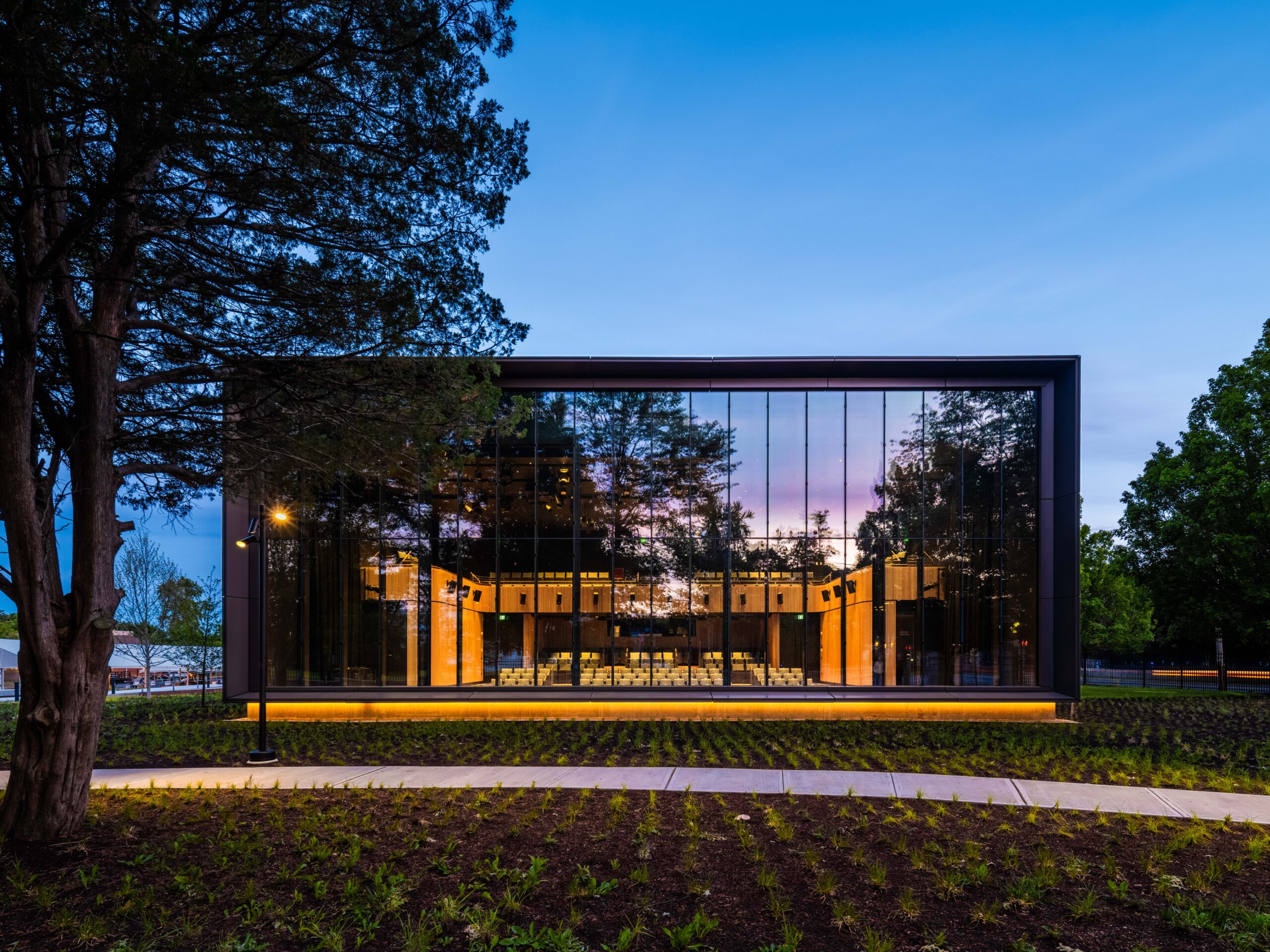 Modern glass building with illuminated interior, reflecting trees and sky at dusk, surrounded by landscaped grounds and a concrete walkway.