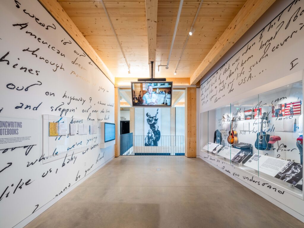 Museum exhibit with handwritten lyrics on the walls, display cases with musical artifacts, and a central video screen above photographs and memorabilia.