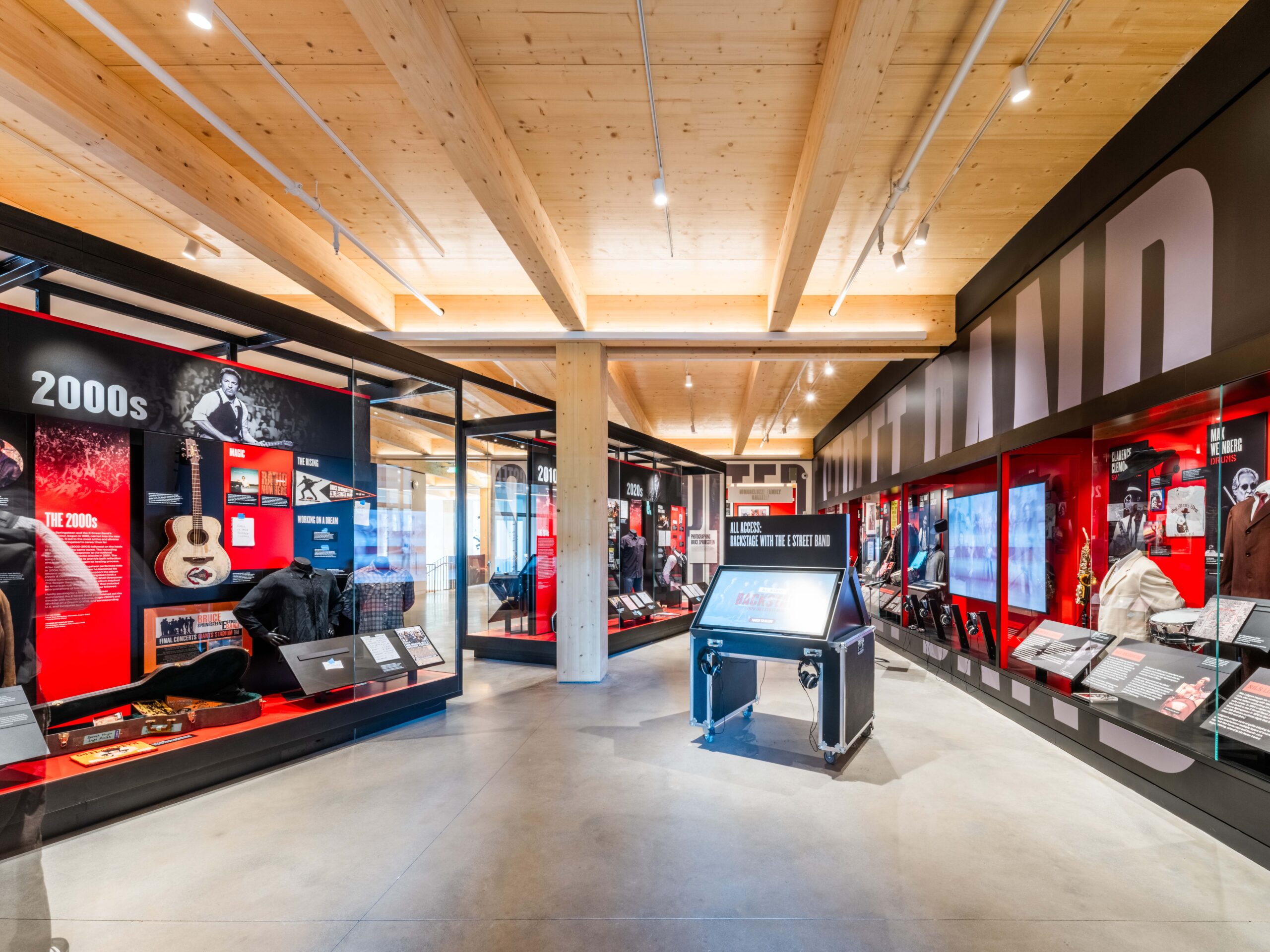 Modern museum exhibit with red and black displays, artifacts, and informational panels focused on music history, set in a spacious room with wood beams and polished concrete floor.