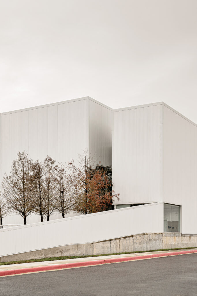 A modern, minimalist white building with clean lines, reminiscent of Neri&Hu's signature style, stands adjacent to a row of thin trees and a concrete ramp under an overcast sky.