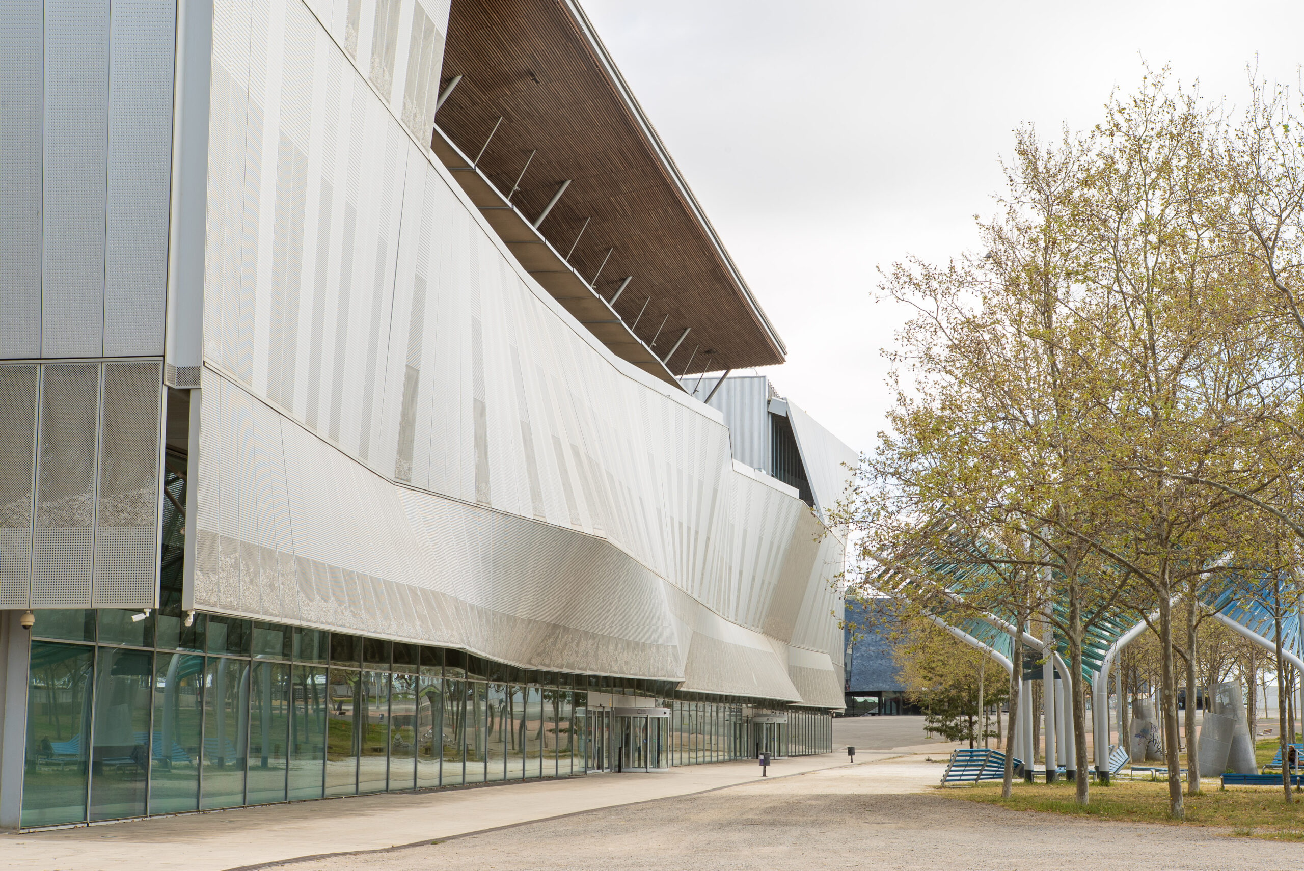 Modern building with a wavy metal façade and large glass windows, surrounded by trees with sparse leaves on a gravel path—an architectural highlight featured at 3daysofdesign.