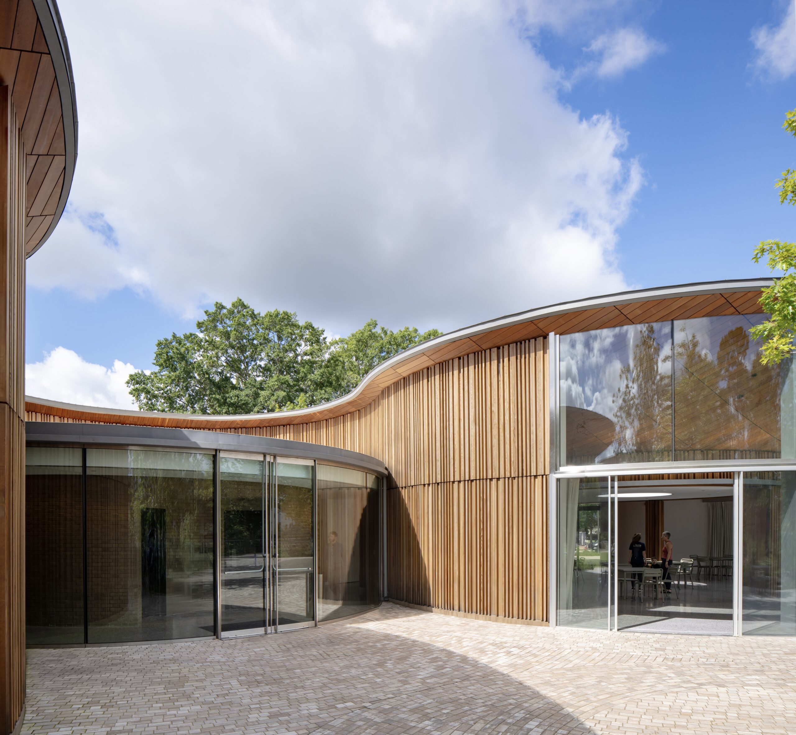 Curved modern building with tall glass walls and vertical wooden slats, featuring an open entrance and a small group of people visible inside—possibly participants at a Camp for the Blind. Blue sky with clouds overhead.