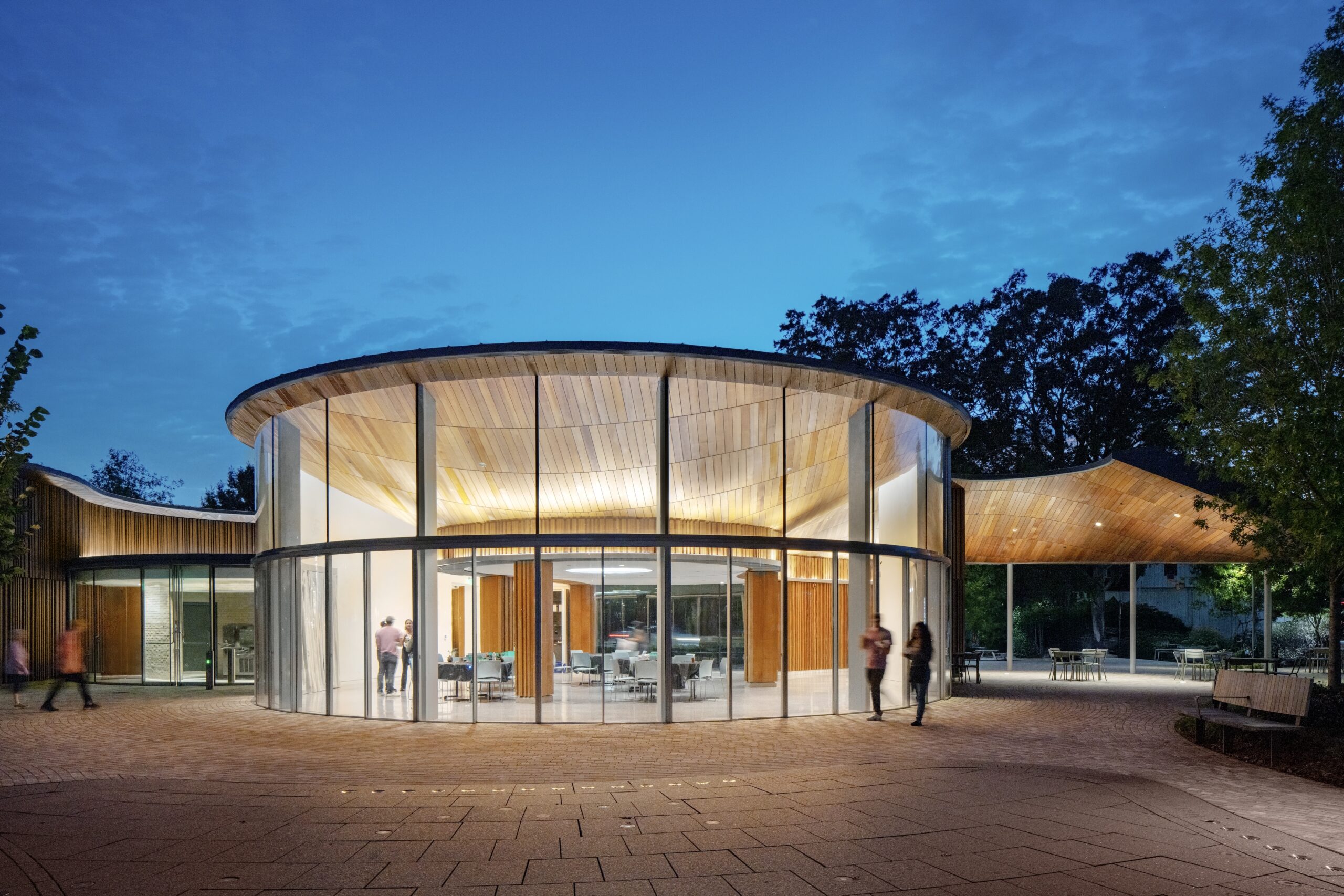 Modern circular glass building with wooden roof, illuminated at dusk. Several people walk nearby on a paved area surrounded by trees, highlighting the welcoming atmosphere of this Camp for the Blind.