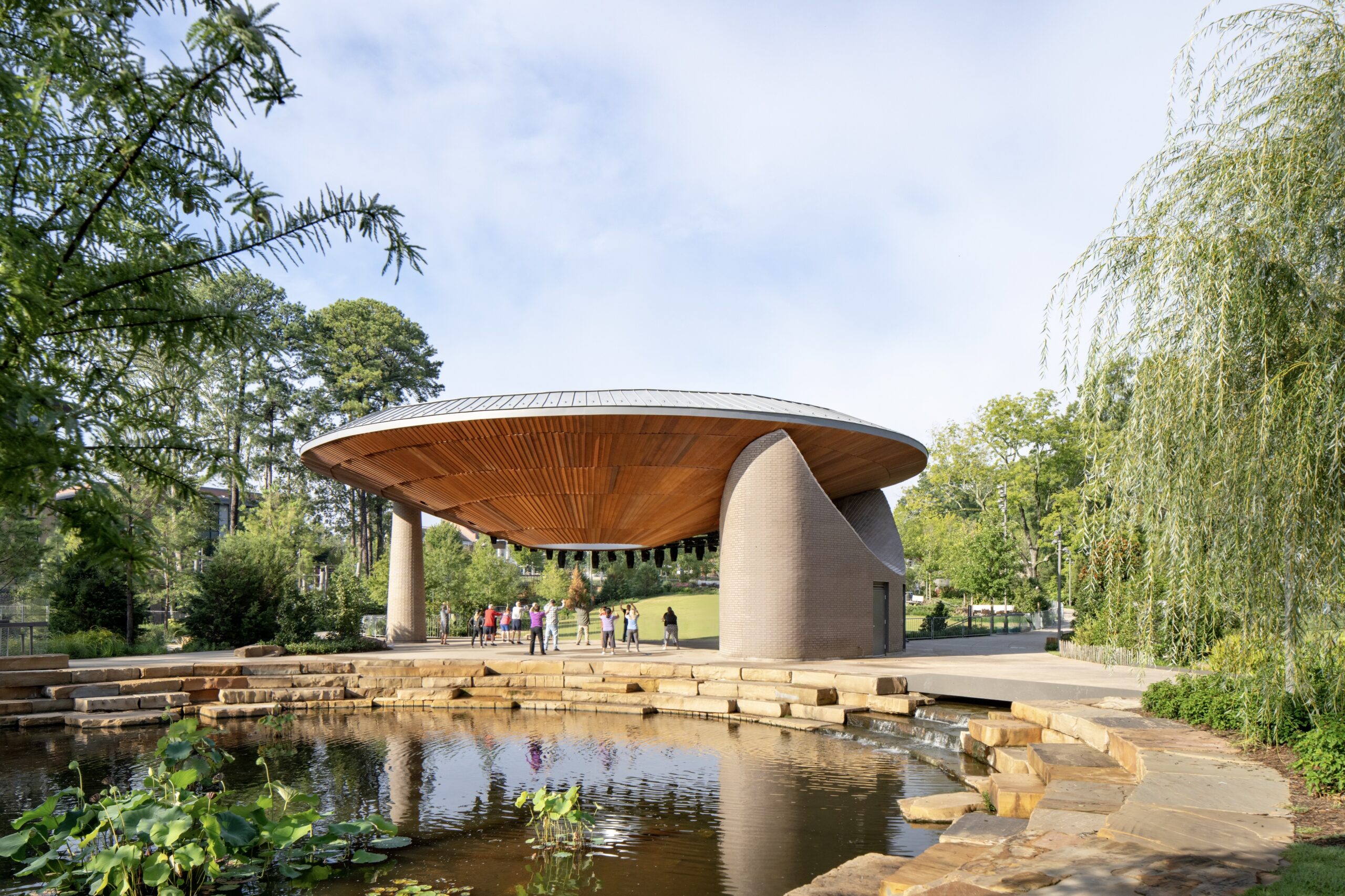 A round, wood-roofed pavilion stands near a pond with stone steps, surrounded by greenery at the Camp for the Blind. Several people are gathered under the pavilion on a sunny day.