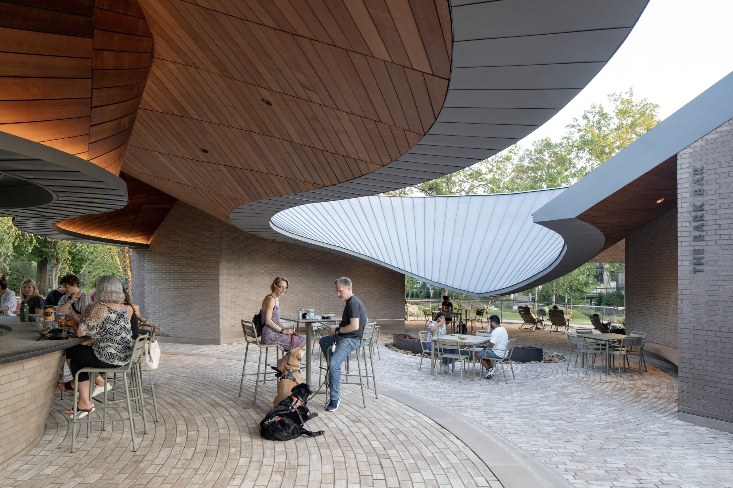 People sit and converse at outdoor tables under a modern, curved wooden pavilion with a bright white canopy—a welcoming atmosphere reminiscent of gatherings at Camp for the Blind. A dog lies on the ground beside a couple at the center, suggesting the inclusive spirit so often fostered in such community spaces.