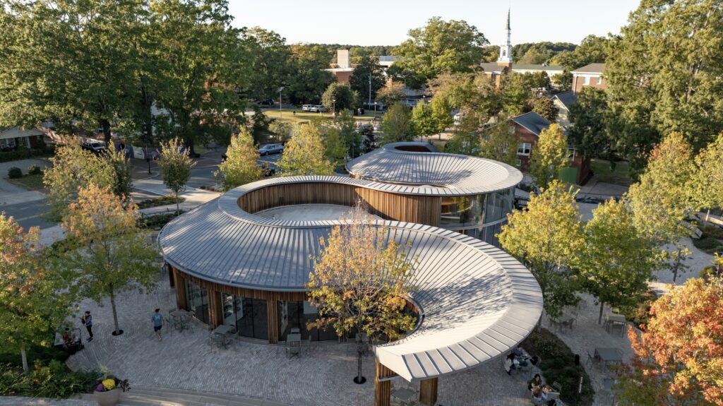 Curved metal-roofed modern buildings surrounded by trees, with people walking on stone paths in a suburban neighborhood—a peaceful setting reminiscent of a thoughtfully designed Camp for the Blind.