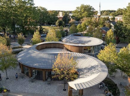 Curved metal-roofed modern buildings surrounded by trees, with people walking on stone paths in a suburban neighborhood—a peaceful setting reminiscent of a thoughtfully designed Camp for the Blind.