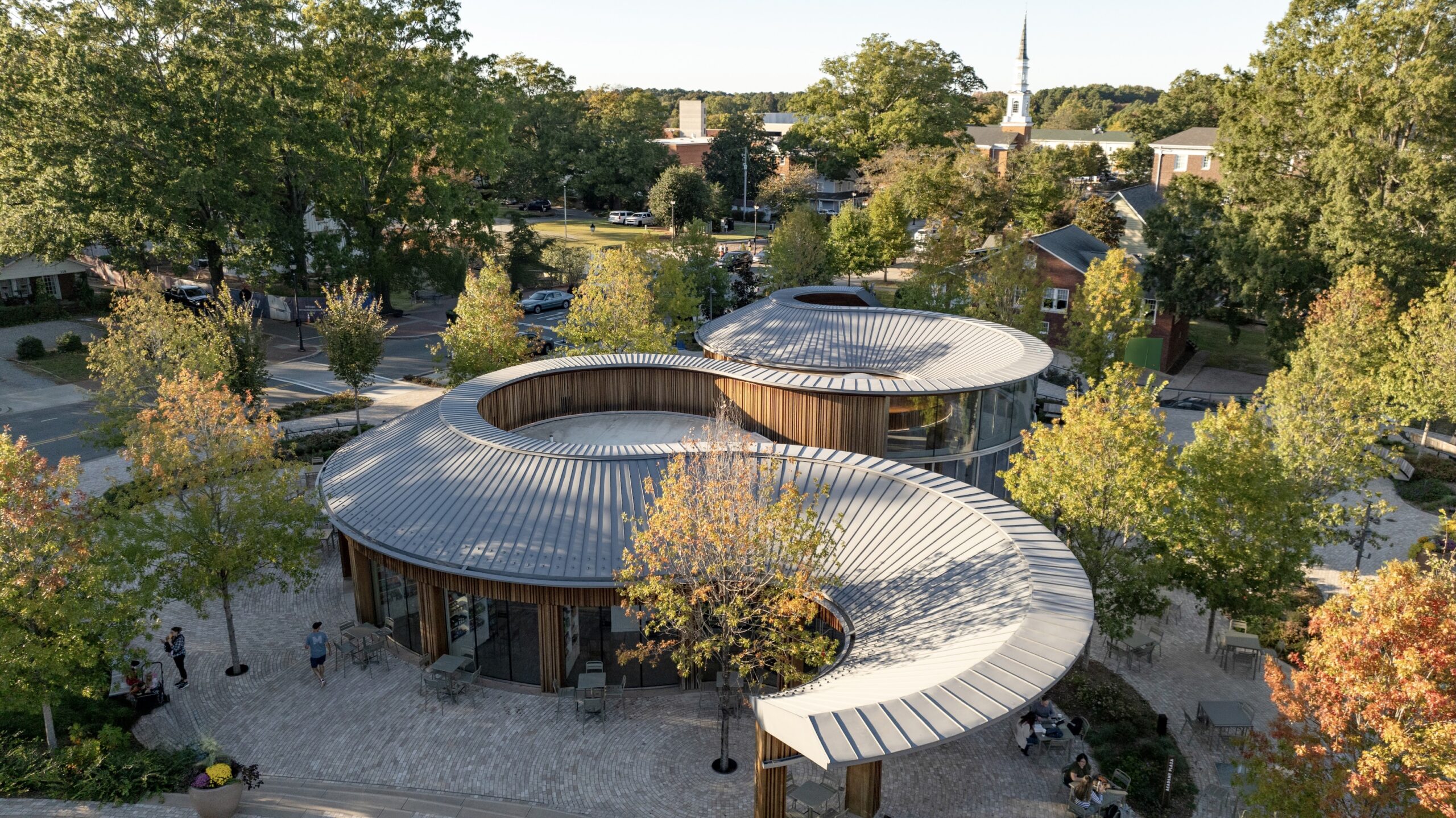 Curved metal-roofed modern buildings surrounded by trees, with people walking on stone paths in a suburban neighborhood—a peaceful setting reminiscent of a thoughtfully designed Camp for the Blind.